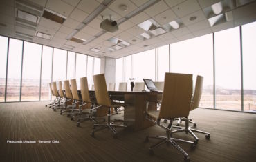 Ein Meetingtisch steht in der Mitte eines großen Büroraums. Darum herum sind weiße Bürostühle gestellt.